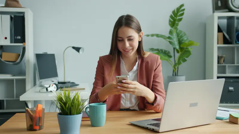 Atendente respondendo mensagens no celular em frente ao notebook